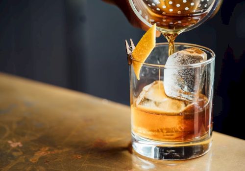A glass of whiskey with ice on a rustic bar, poured from a decanter, garnished with an orange twist.