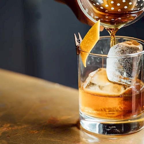 A glass of whiskey with ice on a rustic bar, poured from a decanter, garnished with an orange twist.