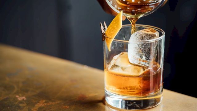 A glass of whiskey with ice on a rustic bar, poured from a decanter, garnished with an orange twist.
