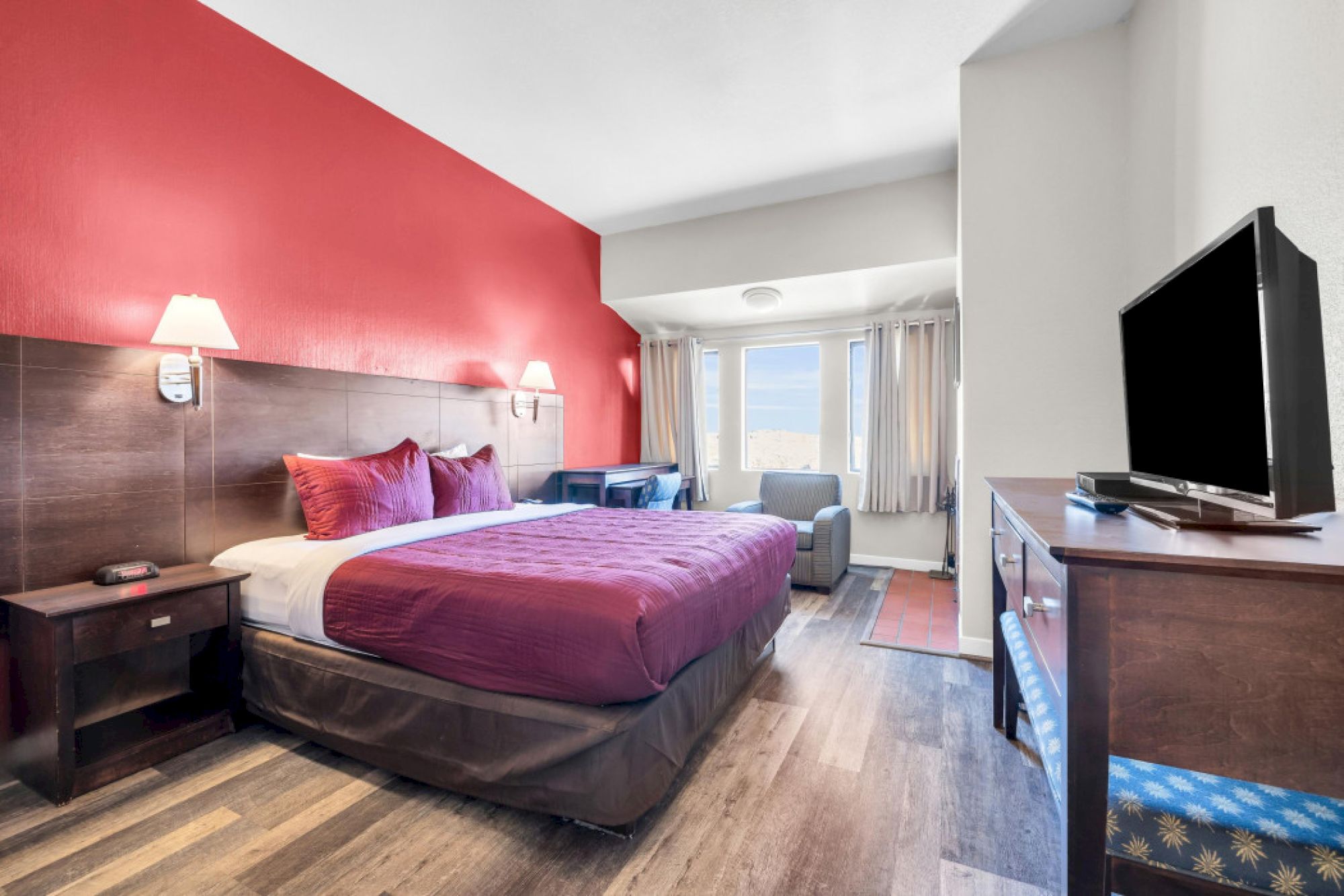 A hotel room with a red accent wall, bed, TV, armchair, and window with curtains.