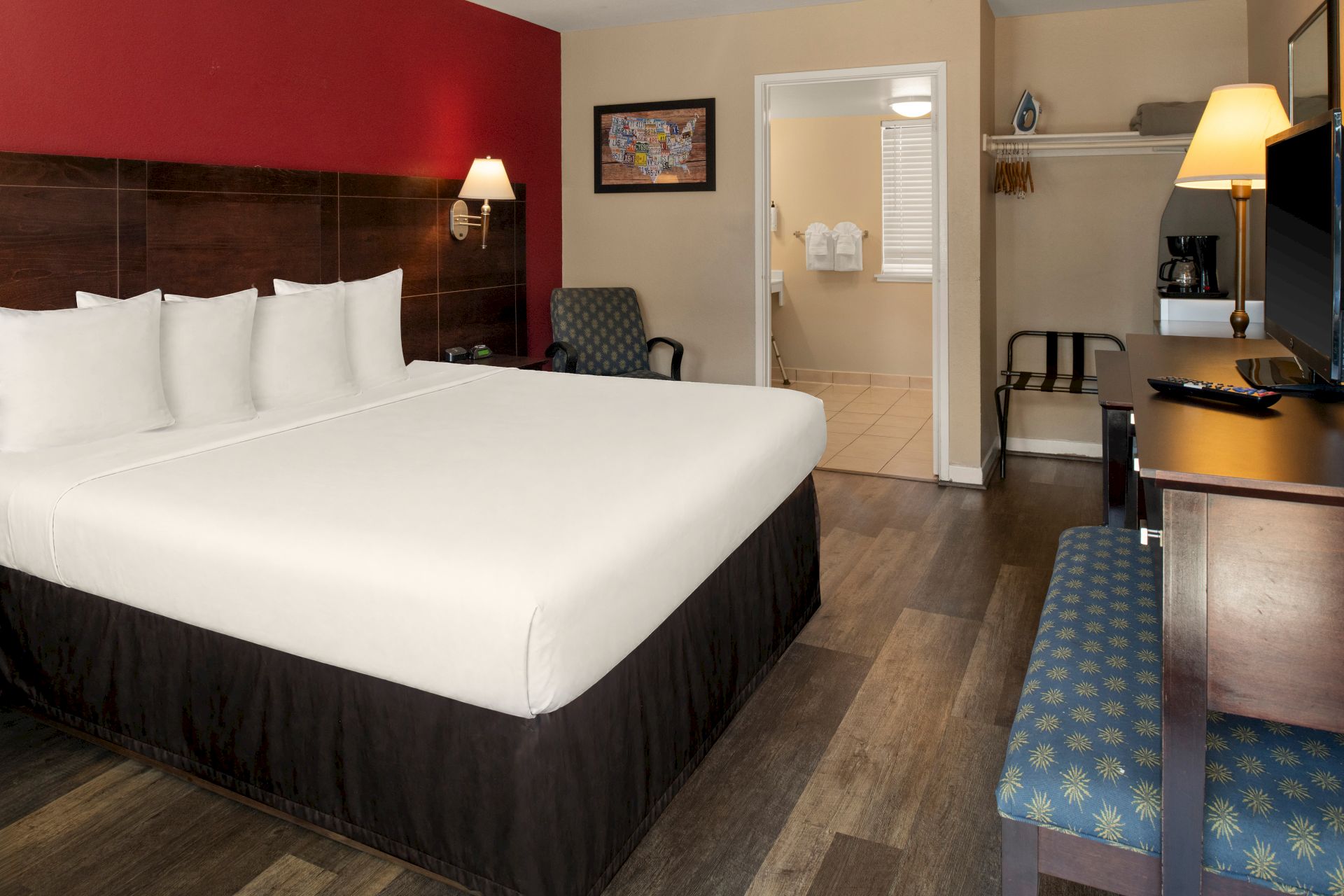 Hotel room with a large bed, red accent wall, desk, TV, seating area, and a hallway leading to a bathroom; tidy and modern.