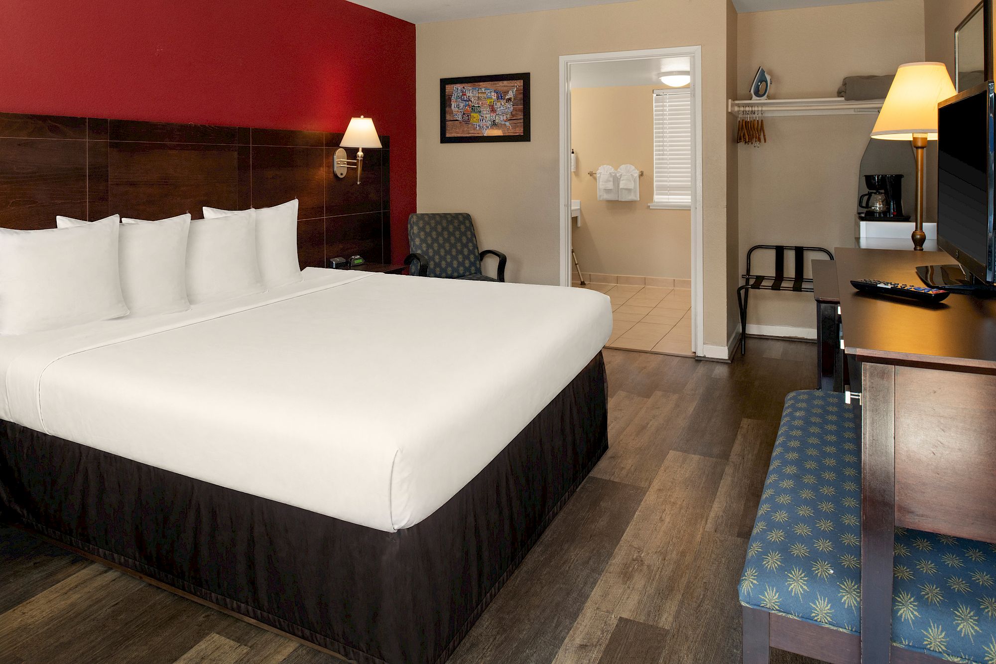 Hotel room with a large bed, red accent wall, desk, TV, seating area, and a hallway leading to a bathroom; tidy and modern.