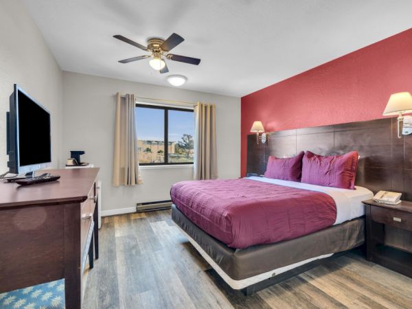 A cozy hotel room with a double bed, red accents, wall lamps, TV, window, and wooden floor, creating a welcoming atmosphere.