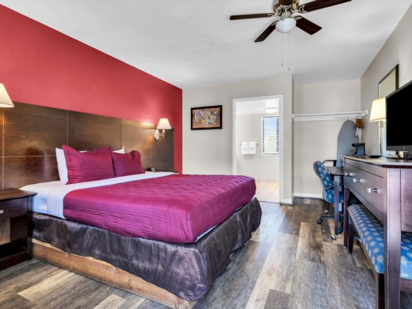 The image shows a hotel room with a bed, TV, desk, chair, ceiling fan, and a red accent wall.