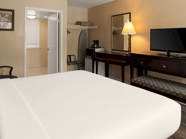 A tidy hotel room with a large bed in the foreground, a desk with a lamp, TV, and bench; open closet, chair, and a doorway to a bathroom&mdash;bright and simple.