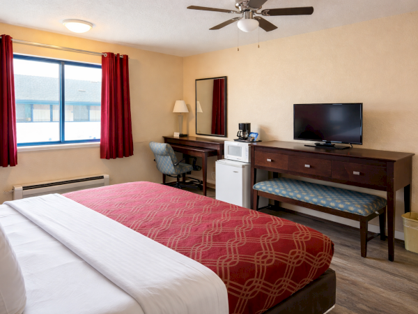 A hotel room with two queen beds, red patterned blankets, a desk, chair, flat-screen TV, mini-fridge, microwave, and a window with red curtains.