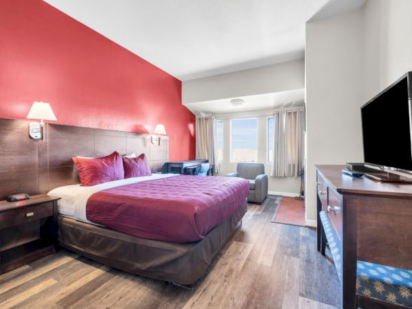 A hotel room with a red accent wall, bed, TV, and seating area by the window, combining modern decor with comfort.
