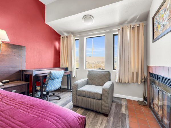 A cozy room with a bed, desk, chair, and fireplace. Bright red accent wall and two windows with curtains let in light, enhancing comfort.