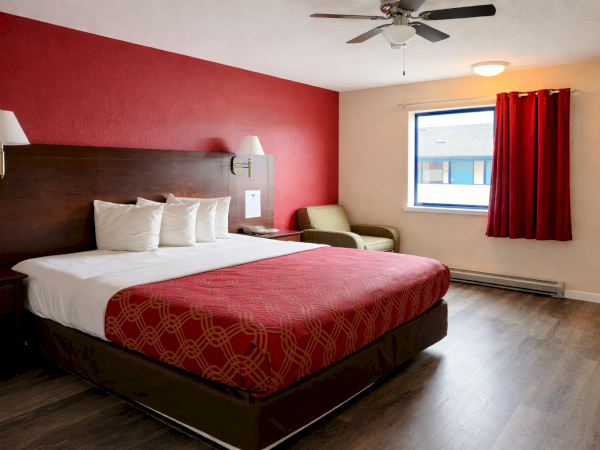 A hotel room with a large bed, red accent wall, two nightstands, ceiling fan, window with red curtains, and a small chair in a bright, simple setup.