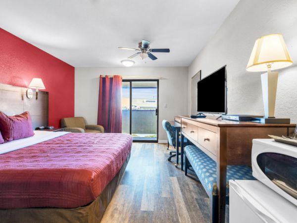 A hotel room featuring a large bed, TV, desk, microwave, and a view through sliding glass doors, with a red accent wall and modern decor.