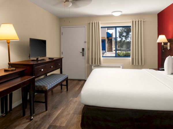 A tidy hotel room with a large bed, desk, TV, and window view; warm lamps, neutral tones, and a red accent wall. Top it at 140 characters, always ending the sentence.