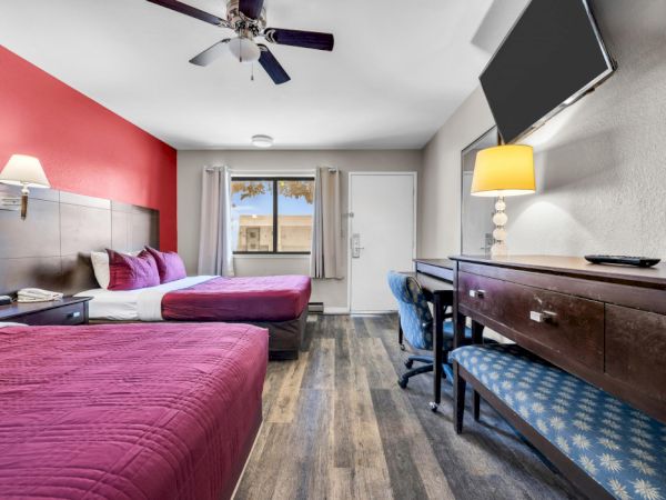 The image shows a hotel room with two beds, a desk, a wall-mounted TV, a lamp, and a window with curtains.