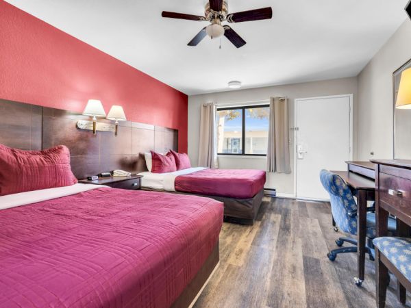 A hotel room with two beds, red accents, a desk, chair, lamp, and ceiling fan, with a window and door visible.