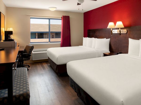 A hotel room with two double beds, red accent wall, desk and chair, flat-screen TV, window with a view, and warm lighting.