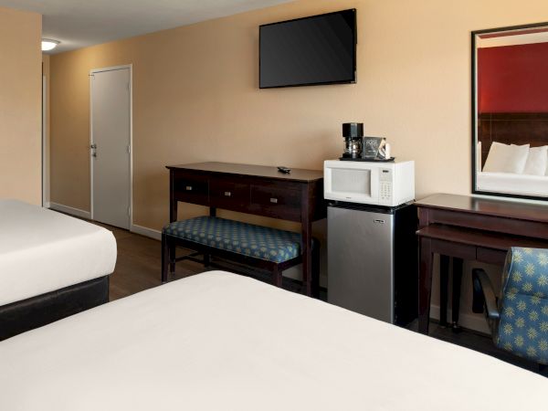 A hotel room with two beds, a desk and bench, a wall-mounted TV, a mini-fridge with a microwave, and a mirror above a counter.