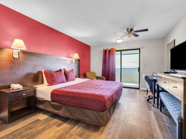 The image shows a hotel room with a bed, TV, desk, chair, and window with a view, featuring red and brown decor.