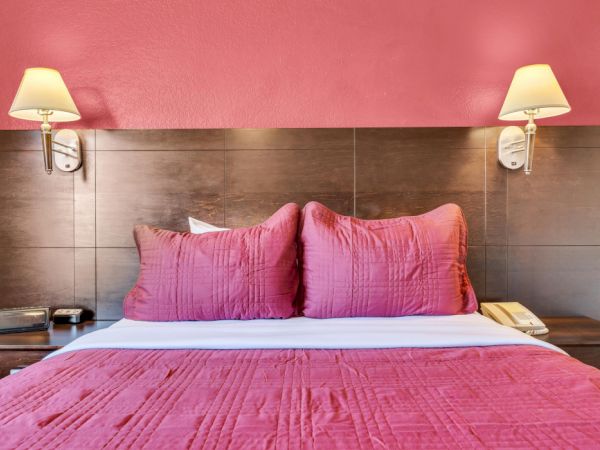 A bed with pink bedding, two pillows, and wall lamps in a room with a pink wall, along with a phone and a clock on the bedside table.