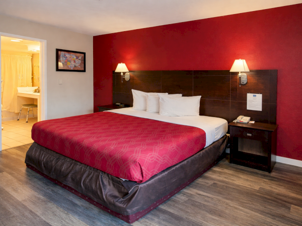 A hotel room with a red accent wall, king bed, white linens, two wall lamps, dark wood headboard, and an open door to a bathroom.