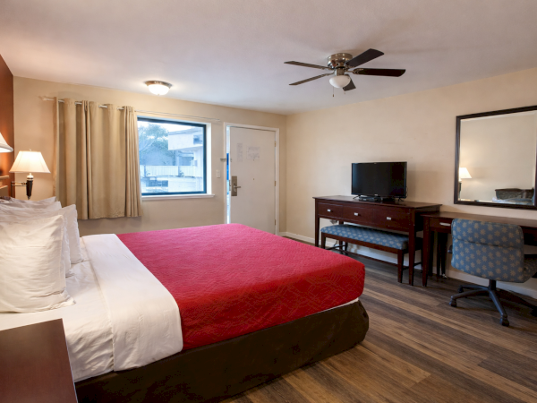 A hotel room with a king bed, red comforter, white sheets, nightstands, a desk with a chair, a wall mirror, a flat-screen TV, window with curtains, and a ceiling fan.