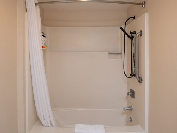 A small bathroom with a bathtub/shower combo, white tile surround, a white shower curtain, wall-mounted showerhead, and a towel at the tub edge.