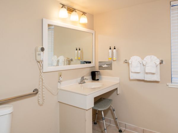 A small, clean bathroom with a white sink and mirror, wall-mounted hair dryer, two shelves with toiletries, a stool, towels, and a window with blinds.