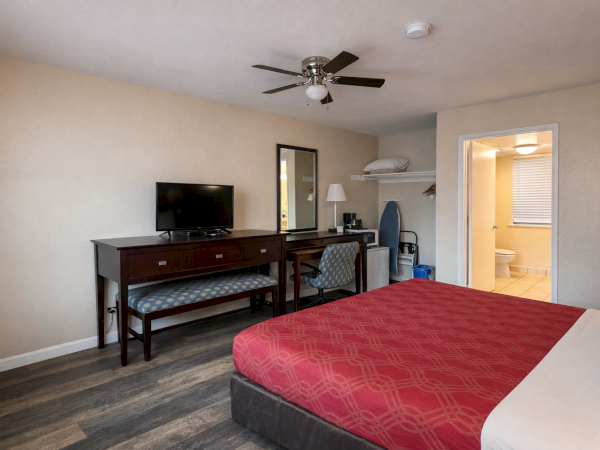 A hotel-like bedroom with a red duvet, dark wood desk and chair, small TV, ceiling fan, and an open doorway to a bathroom; simple and tidy.