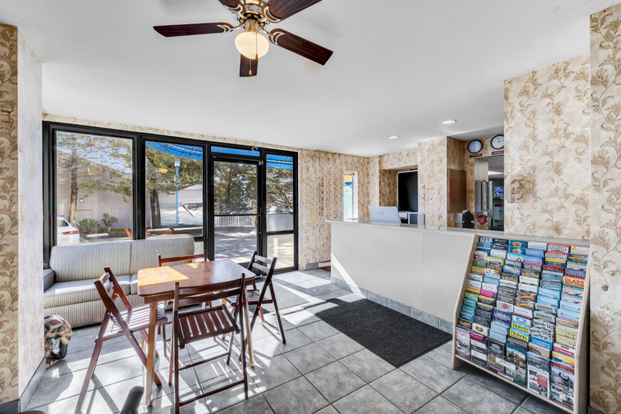 A small lobby with a table, chairs, brochures on a stand, and a reception desk, featuring large windows and a ceiling fan.