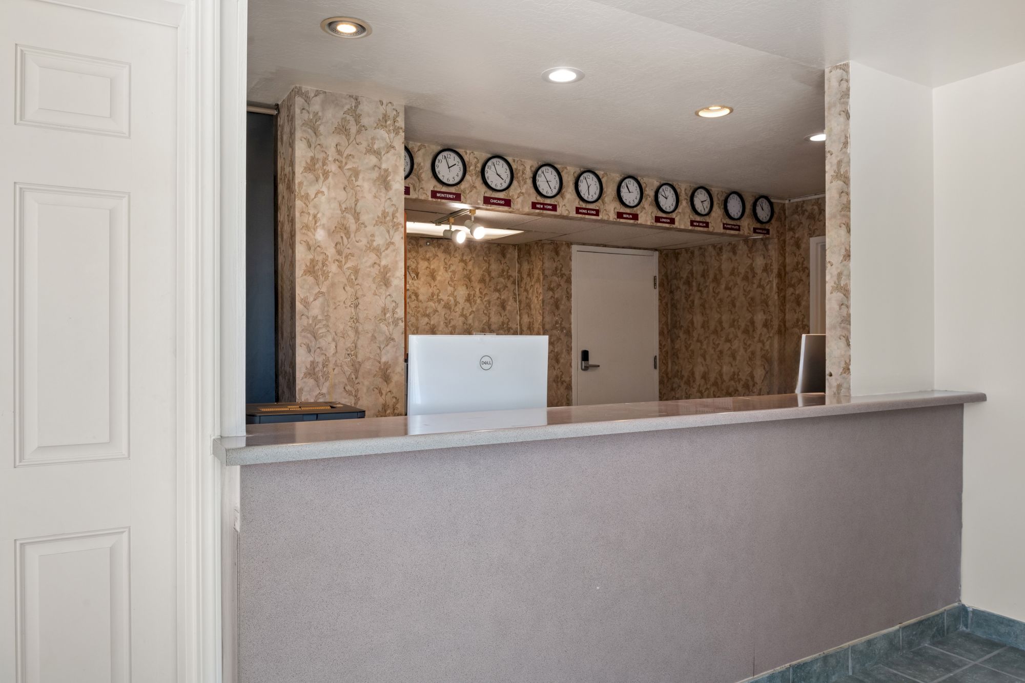 A simple hotel front desk area with a long counter, wallpapered walls, clocks displayed on the back wall, and a doorway behind the counter.