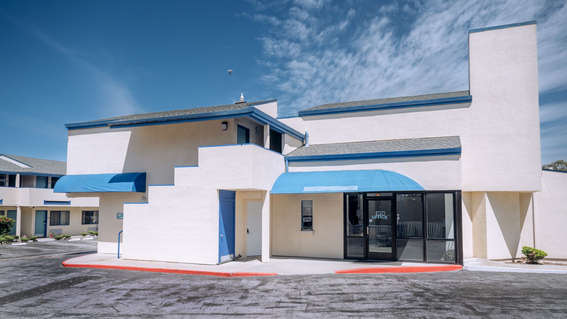 A modern, white building with blue accents and angular, stepped design; glass entry, small awnings, and a red curb in front.