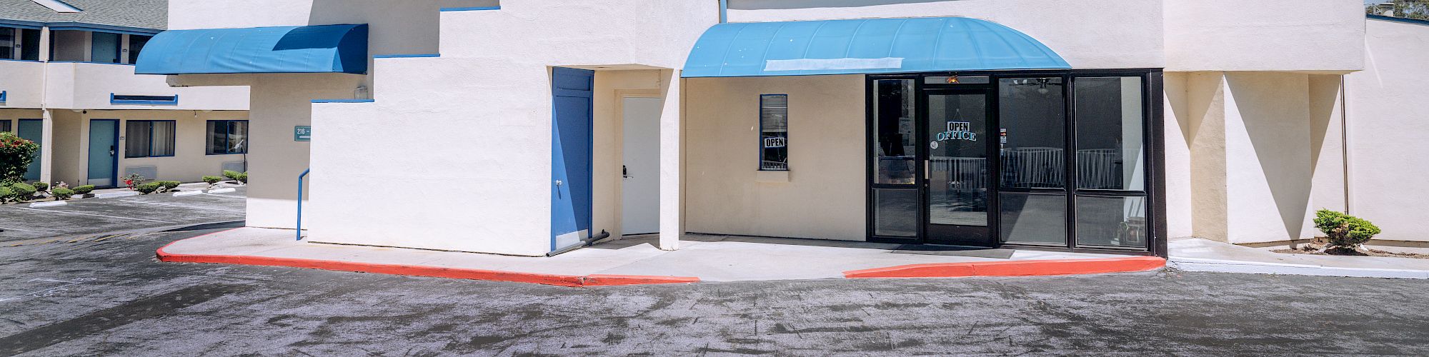 A modern, white building with blue accents and angular, stepped design; glass entry, small awnings, and a red curb in front.