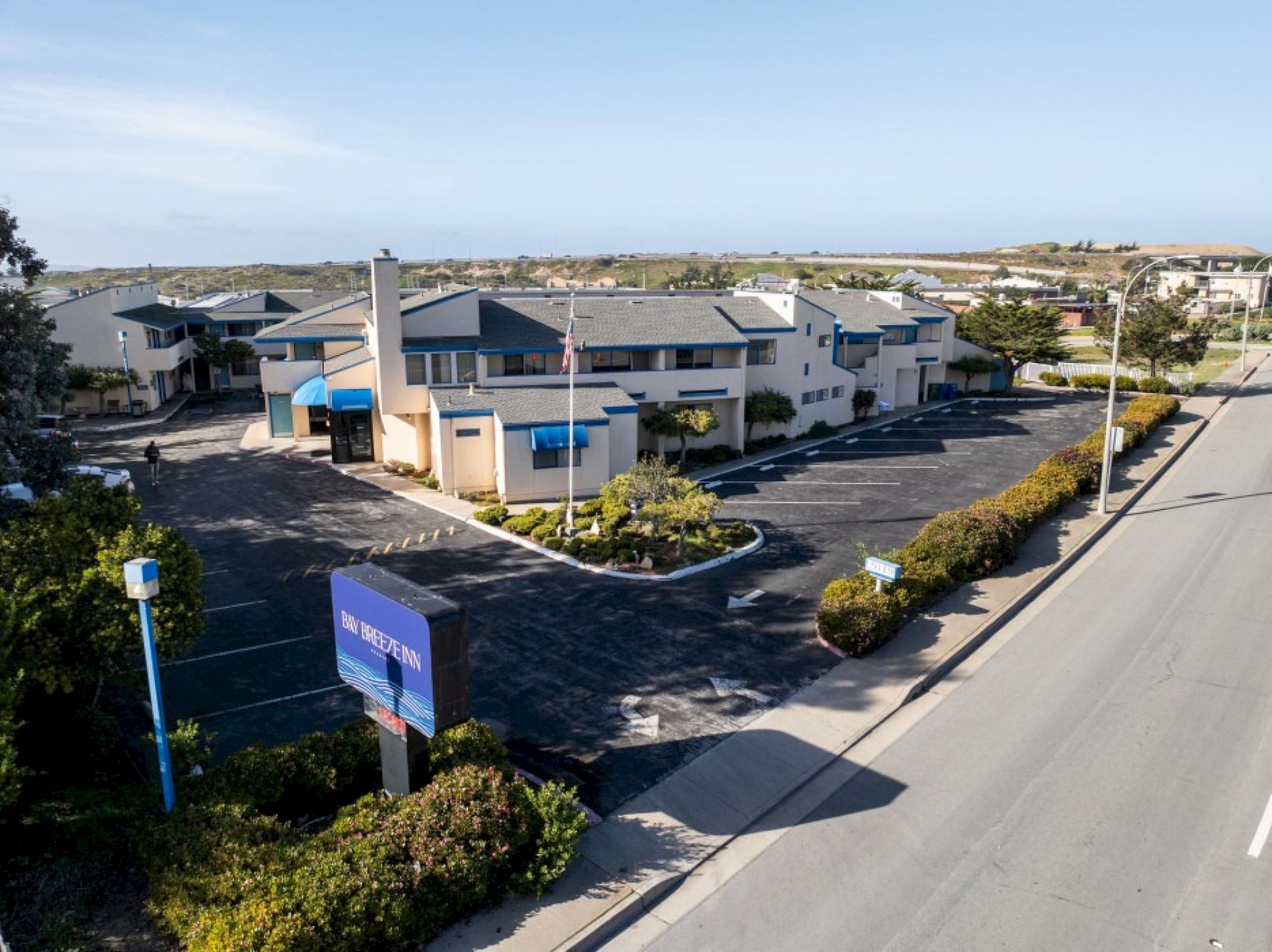 This image shows a two-story building with a parking lot, surrounded by trees and shrubs, adjacent to an empty street under a clear sky.