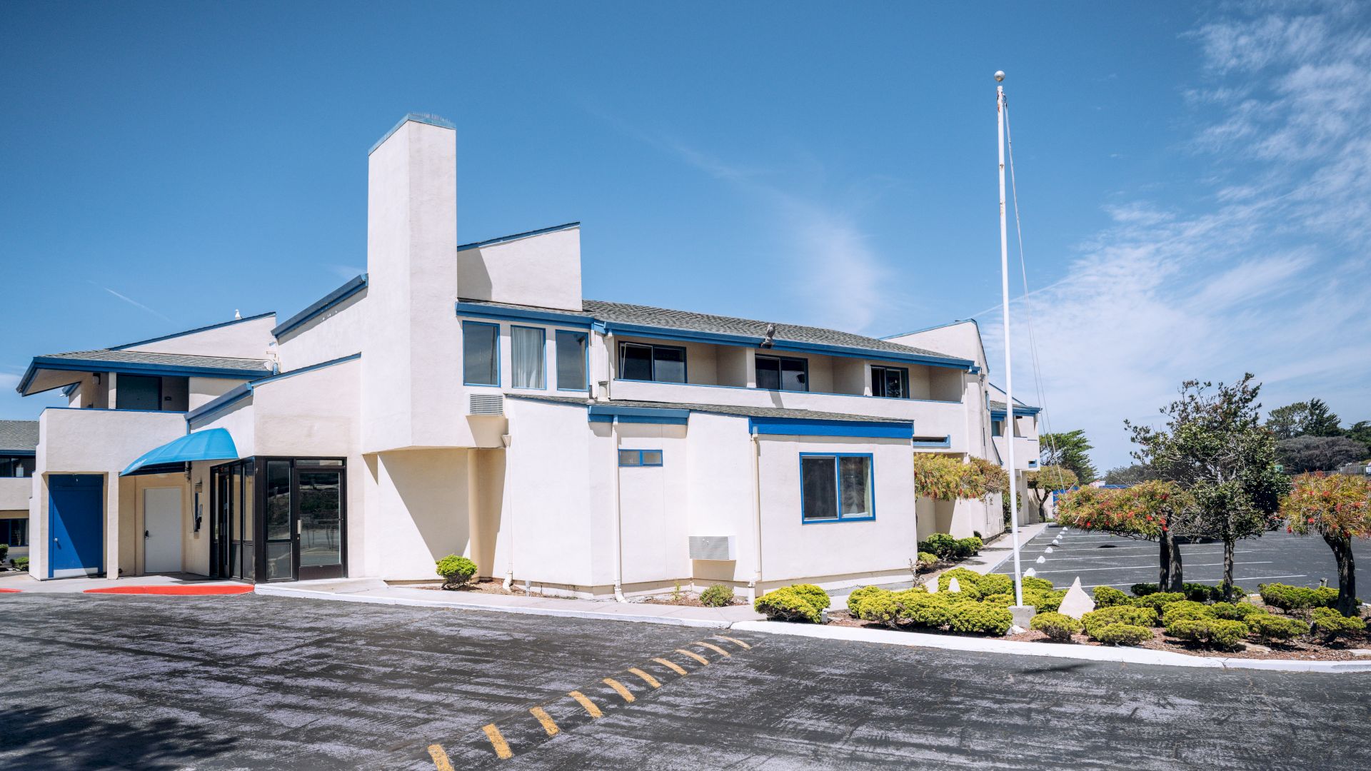 A white two-story motel with blue trim, a tall chimney, a glass entry, small shrubs, and a parking lot under a clear blue sky, end.