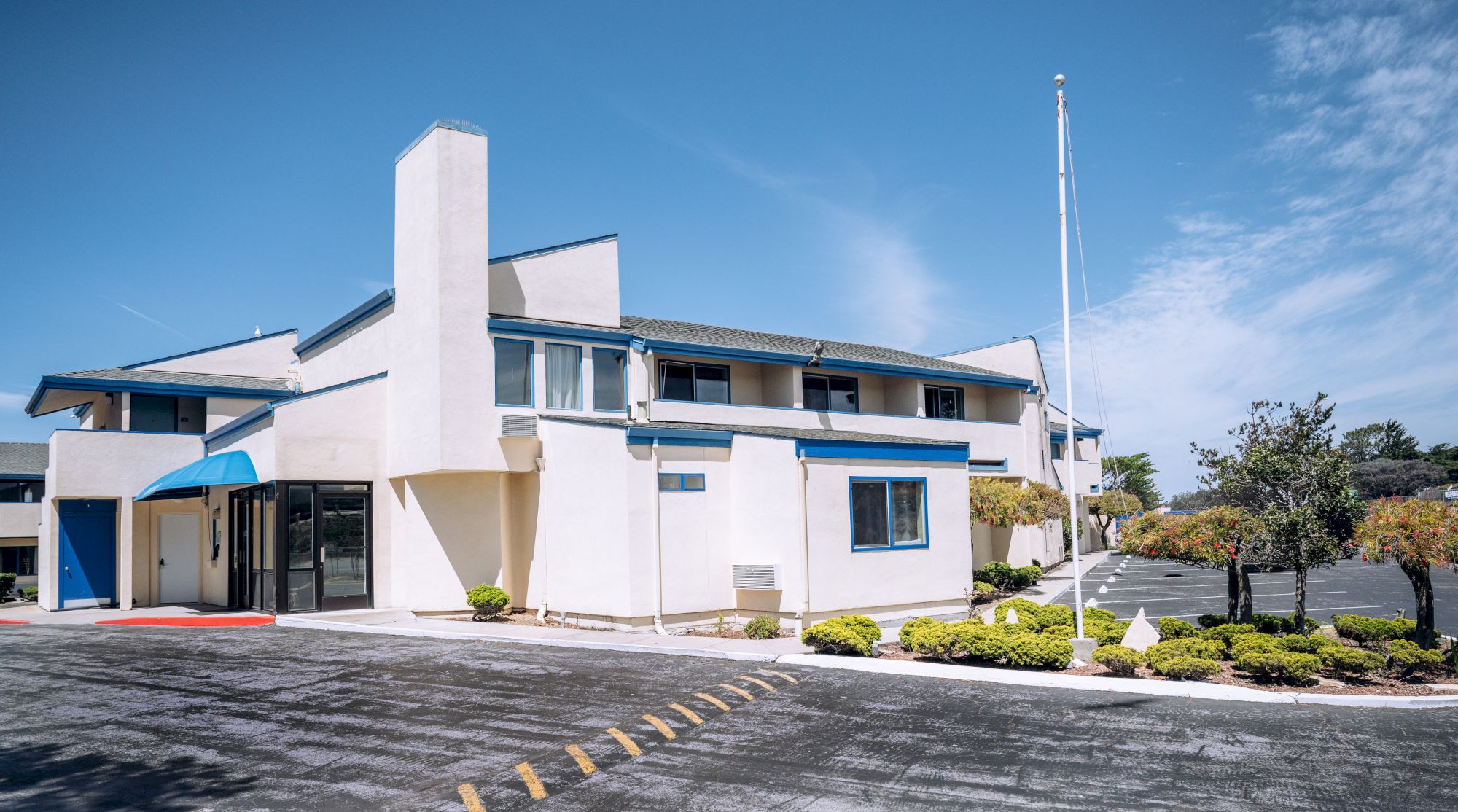 A white two-story motel with blue trim, a tall chimney, a glass entry, small shrubs, and a parking lot under a clear blue sky, end.