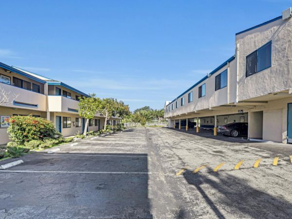 The image shows a paved driveway between two apartment buildings with covered parking on the right and some greenery on the left.