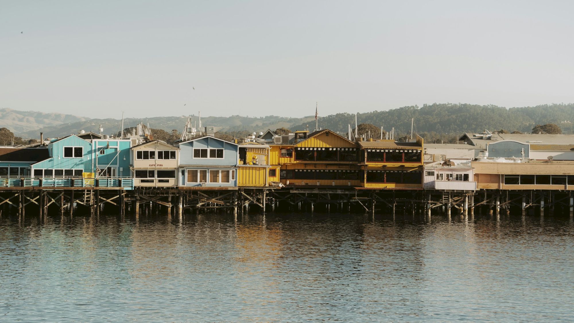 A row of colorful buildings on a waterfront, with mountains in the background, reflecting in the calm water below.