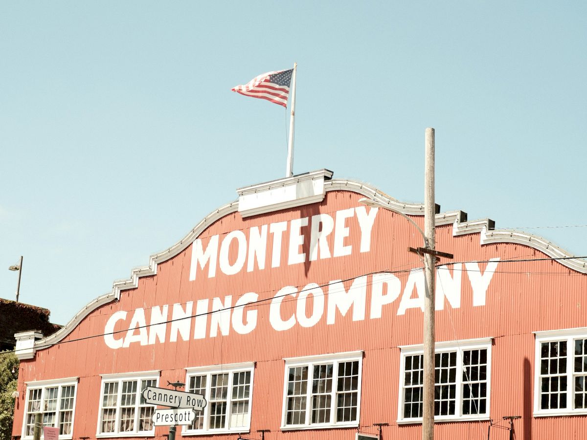A red building with "Monterey Canning Company" on it, an American flag on top, and cars parked along the street.