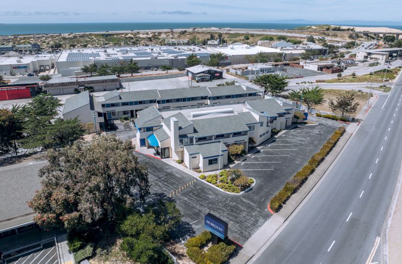 An aerial view of a small commercial area along a highway, with office buildings, parking lots, a few trees, and a road running parallel to the row of shops.