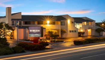 A modern office building at dusk with warm lights, a blue sky, and a sign reading &ldquo;Bay Breeze&rdquo; by a lit driveway.