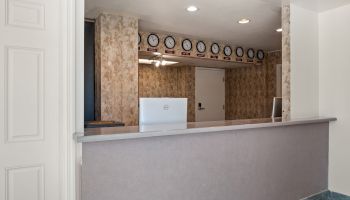 A reception desk in a small lobby with floral wallpaper, clocks along the back wall, ceiling lights, and a white counter facing the entry.