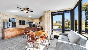 The image shows a room with a wooden table, chairs, a sofa, large windows, ceiling fan, and a counter area with appliances and cabinets.