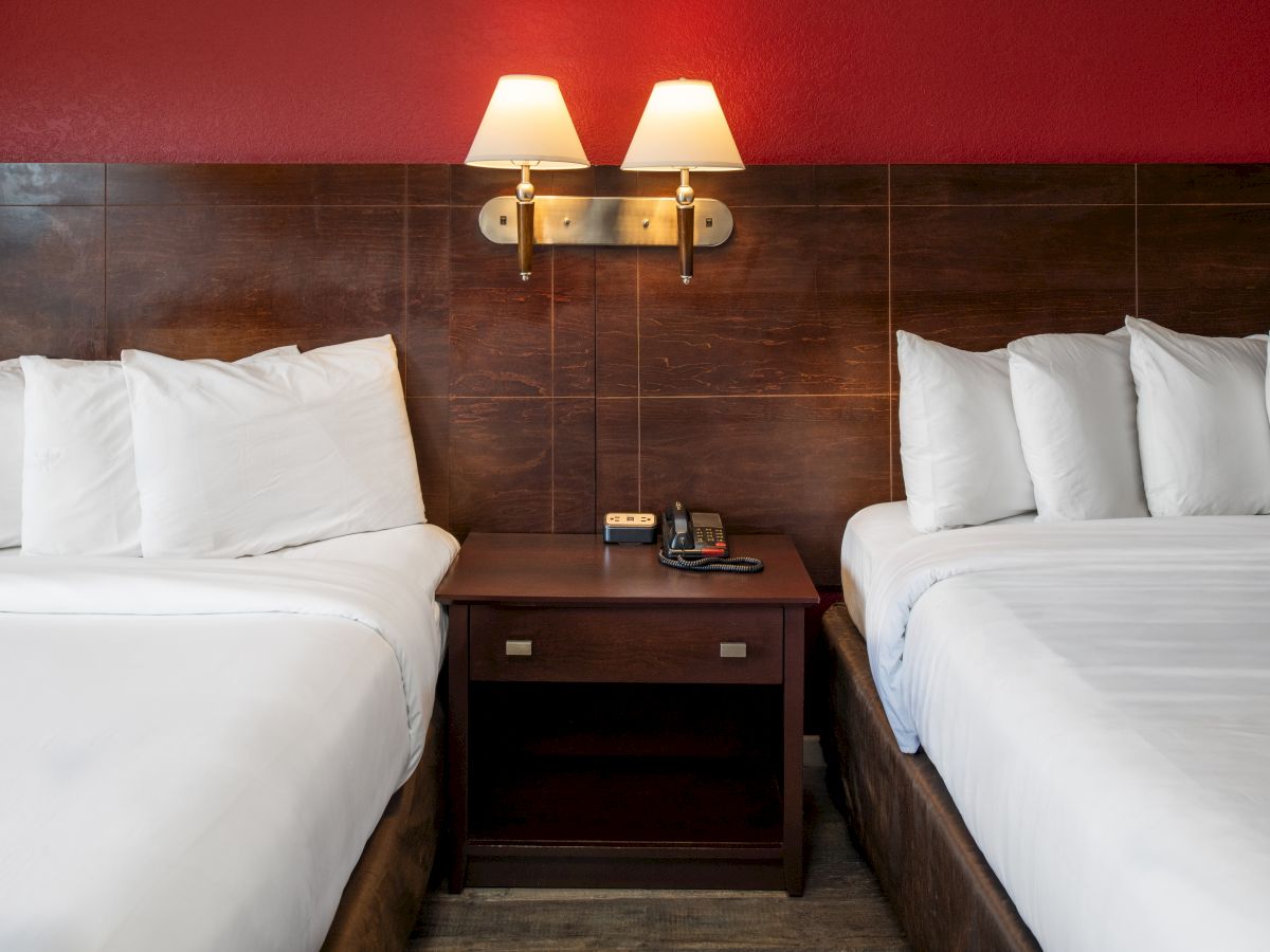 Two queen beds with white linens, a shared nightstand, wall lamps, and a red accent wall behind them.