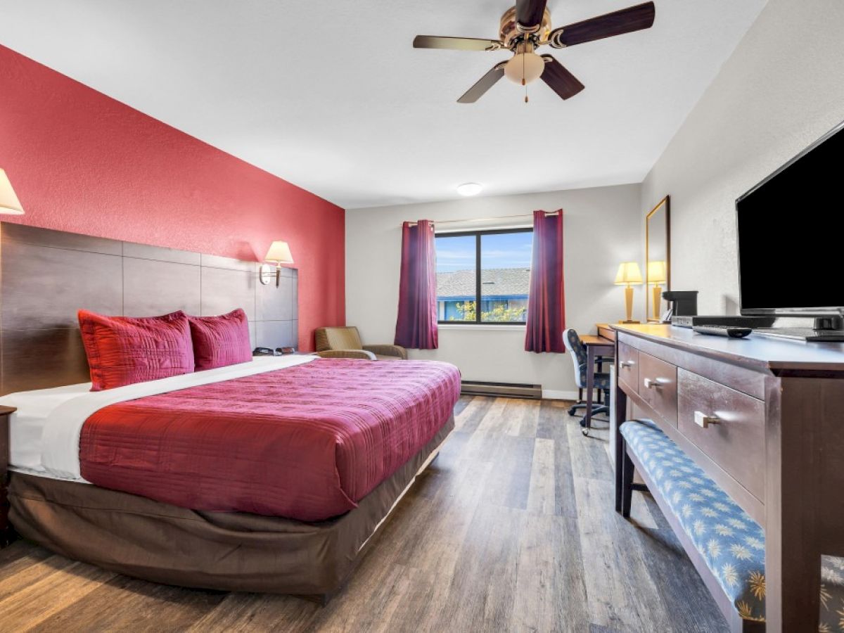 The image shows a hotel room with a large bed, TV, desk, chair, and window with curtains. The decor is warm with a red accent wall.
