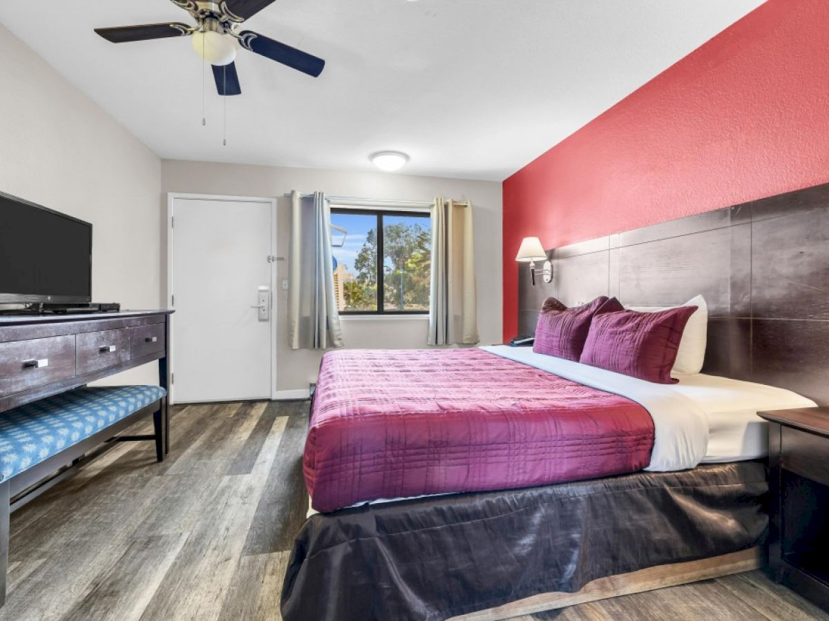 The image shows a hotel room with a double bed, red accent wall, TV, two nightstands, and a window with curtains.