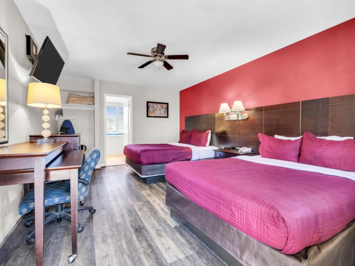 The image shows a hotel room with two beds against a red accent wall, a desk, chair, lamps, and a wall-mounted TV.