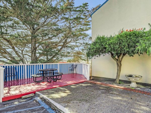 A patio with a red floor, picnic table, trees, and a tall white building wall in the background.