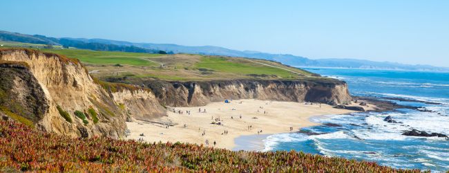 A scenic coastline with cliffs, a sandy beach with people, and the ocean. Lush greenery tops the cliffs under a clear blue sky.