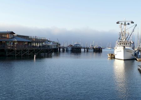A calm harbor scene with a fishing boat docked on the right and buildings on stilts on the left, under a clear blue sky.