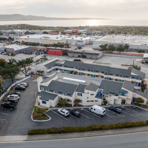 An aerial view of a small business complex with white buildings, parking lots, and a few vehicles, set against a flat, industrial area and distant horizon.