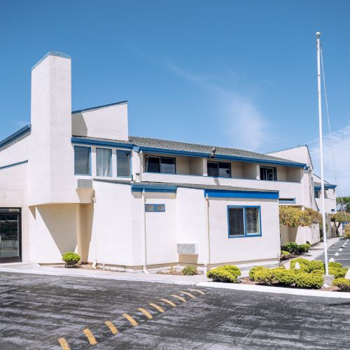 A retro-style two-story motel with white walls, blue trim, a corner office tower, a long front window, and a small parking lot under a clear blue sky.