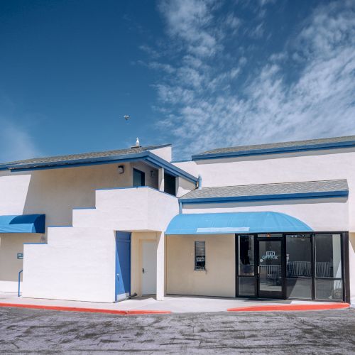 A white, modern building with blue accents and curved awnings; glass entrance, small shrubs, and a clear blue sky above.